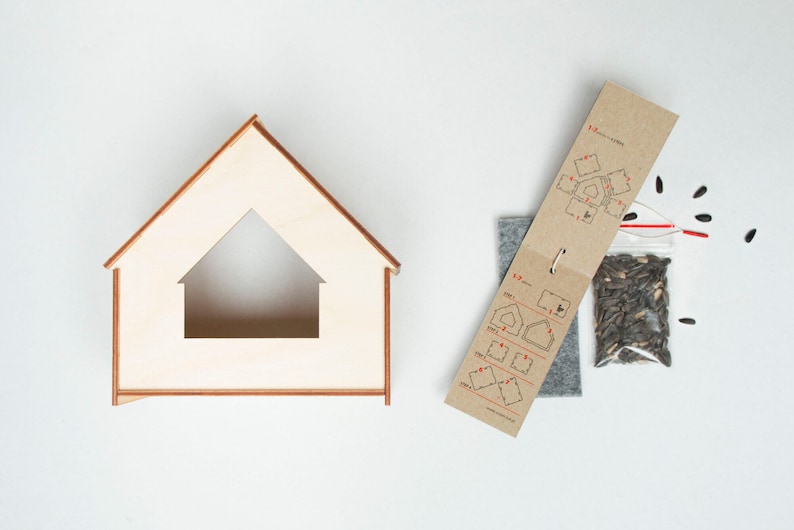 Może przedstawiać: Zestaw drewnianego domku dla ptak&oacute;w z ramą w kształcie domu, paczką nasion słonecznika, szarym kawałkiem filcu i brązową papierową instrukcją. Domek dla ptak&oacute;w ma tr&oacute;jkątny dach i otw&oacute;r w kształcie domu.