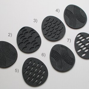May include: Seven dark gray, egg-shaped coasters with various carved designs. The designs include curved lines, curved dashes, and oval cutouts. The coasters are arranged on a white surface, with a studio setup visible in the background.