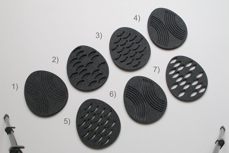 May include: Seven dark gray, egg-shaped coasters with various carved designs. The designs include curved lines, curved dashes, and oval cutouts. The coasters are arranged on a white surface, with a studio setup visible in the background.
