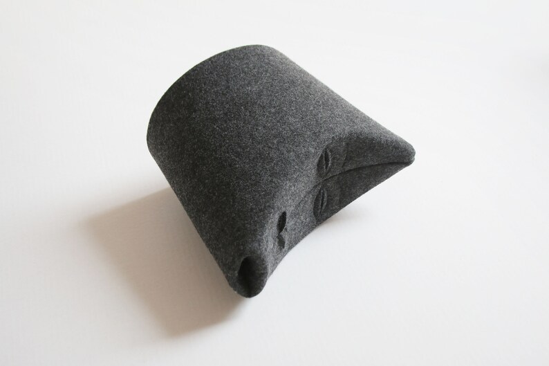 May include: A dark gray felt hat with a unique, angular design. The hat features a cylindrical base that tapers to a pointed end. Three oval-shaped cutouts are visible on the side. The hat is set against a white background.