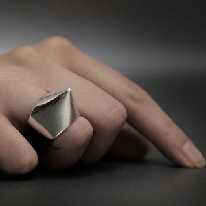 May include: A silver ring with a pointed, triangular design. The ring is worn on a finger.