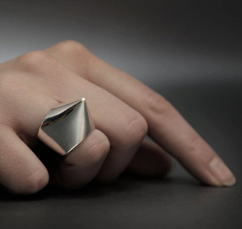 May include: A silver ring with a pointed, triangular design. The ring is worn on a finger.