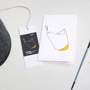 May include: A dark gray felt hat with a tag and a greeting card. The tag and card feature a minimalist illustration of a duck-shaped object with yellow accents. A paintbrush and orange ink bottle are also present.