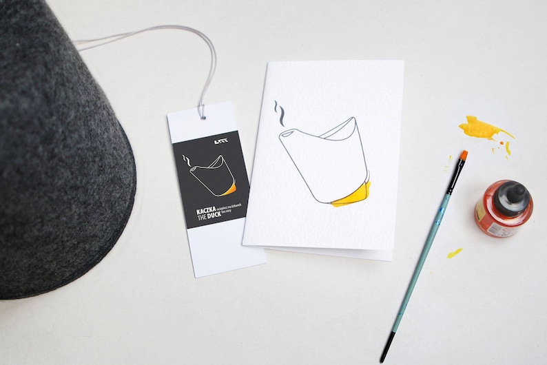 May include: A dark gray felt hat with a tag and a greeting card. The tag and card feature a minimalist illustration of a duck-shaped object with yellow accents. A paintbrush and orange ink bottle are also present.