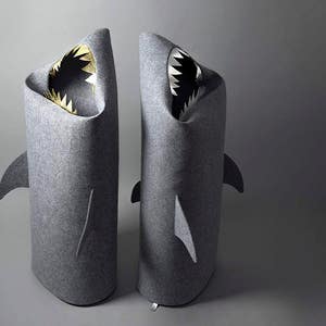 May include: Two gray felt shark-shaped storage bins. Each bin has a wide open mouth with white and black teeth and a gold interior. The bins have fin details on the sides and back, and are designed to stand upright.