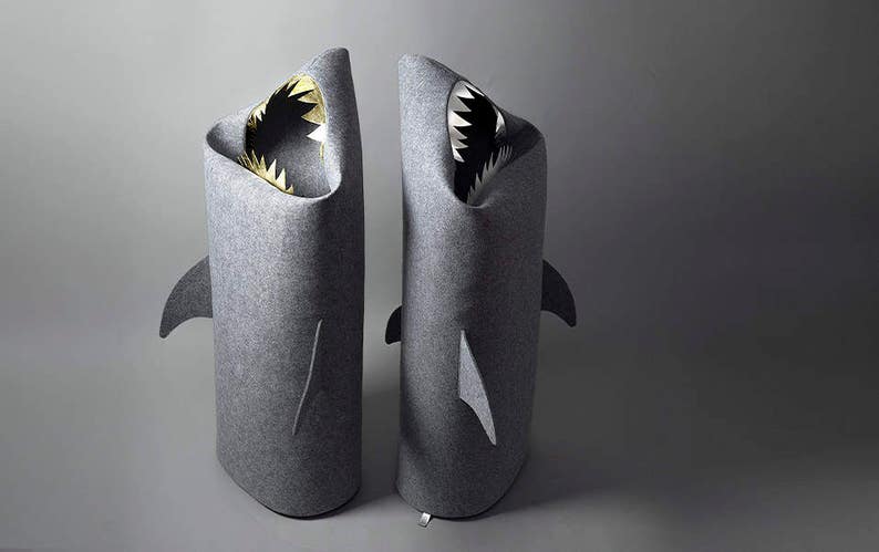 May include: Two gray felt shark-shaped storage bins. Each bin has a wide open mouth with white and black teeth and a gold interior. The bins have fin details on the sides and back, and are designed to stand upright.