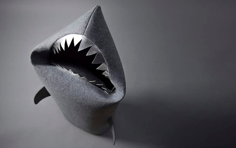 May include: A gray shark-shaped decorative item with a wide-open mouth revealing metallic, jagged teeth. The shark has a pointed fin and a dark gray background. The item is likely made of felt or a similar fabric.