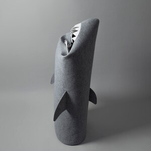 May include: A gray felt shark-shaped bed for pets. The bed has a cylindrical body with fins and a wide open mouth with white teeth. The interior is visible, suggesting a cozy space for a small animal.