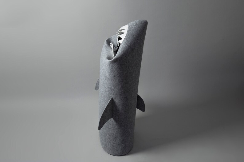 May include: A gray felt shark-shaped bed for pets. The bed has a cylindrical body with fins and a wide open mouth with white teeth. The interior is visible, suggesting a cozy space for a small animal.