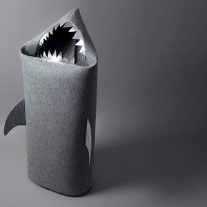 May include: A gray shark-shaped storage bin. The bin is made of a soft, felt-like material and features a shark fin and a mouth with sharp, white teeth. The interior is visible through the open mouth.