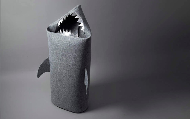 May include: A gray shark-shaped storage bin. The bin is made of a soft, felt-like material and features a shark fin and a mouth with sharp, white teeth. The interior is visible through the open mouth.