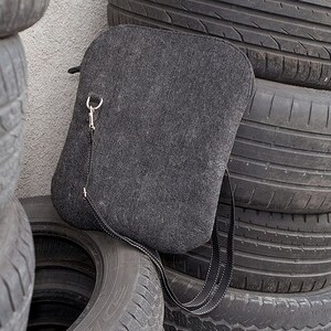 May include: A gray felt crossbody bag with a silver metal clasp and a black adjustable strap. The bag is sitting on a stack of used tires.