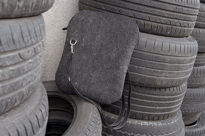 May include: A gray felt crossbody bag with a silver metal clasp and a black adjustable strap. The bag is sitting on a stack of used tires.