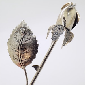 Könnte beinhalten: Eine silberfarbene Metallskulptur in Form einer Rose mit einer geschlossenen Knospe und einem detaillierten Blatt an einem schlanken Stiel. Das Blatt trägt einen 925er Stempel und ein kleines kreisförmiges Emblem aus Sterlingsilber.