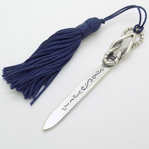 May include: A silver-toned metal bookmark featuring a flat, pointed blade with engraved decorative markings. The top of the bookmark is shaped like a coiled snake and is attached to a dark blue fabric tassel by a thin matching cord.