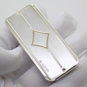 Puede incluir: Un lingote de plata rectangular con un recorte central en forma de diamante y dos líneas verticales que se extienden hacia los extremos redondeados. La superficie muestra pequeños sellos cerca de un extremo, indicando su composición.