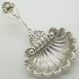 Peut inclure: Une cuillère à thé en argent dotée d'un cuilleron en forme de coquille Saint-Jacques avec des détails cannelés complexes. Le manche est fin et décoratif, se terminant par un petit motif floral au sommet, avec des surfaces métalliques polies.