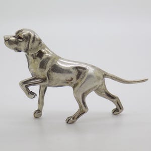 Puede incluir: Una figura de metal en tono plateado de un perro en posición de pie con una pata delantera levantada. La escultura presenta una superficie lisa y reflectante con contornos anatómicos detallados, capturando la postura alerta.
