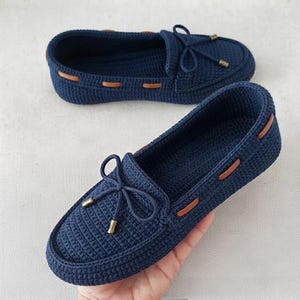 May include: A pair of dark navy blue crochet loafers featuring a textured knit construction, a decorative bow on the vamp, and brown faux leather lacing details threaded through the sides with small metallic gold-toned aglets at the ends.