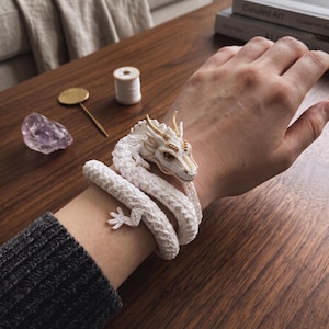 Puede incluir: Una pulsera de dragón tejida a ganchillo en color blanco envuelta alrededor de una muñeca. El dragón presenta intrincadas puntadas de hilo blanco, detalles dorados en los cuernos y el rostro, y pequeños ojos oscuros. El cuerpo largo y enroscado.
