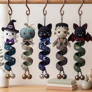 May include: Five hanging crochet ornaments featuring a ghost with a witch hat, a pumpkin, a black cat, a Frankenstein-style figure, and a bat. Each character is attached to a long, spiraling fabric tail decorated with seasonal motifs and bells.