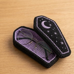 Puede incluir: Un estuche de ganchillo negro con forma de ataúd, interior lavanda y tapa con una luna creciente y estrellas bordadas en color lavanda. Un par de gafas con montura redonda de metal descansa dentro del estuche abierto.