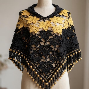 May include: A crochet poncho featuring a black and yellow leaf pattern. The garment is constructed with a series of interconnected maple leaf motifs in black and yellow yarn, finished with a black fringe border accented by small gold-toned beads.