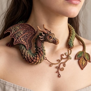 May include: A detailed crochet dragon necklace featuring a textured, multi-coloured body in shades of brown, green, and orange. The dragon has intricate wings, a long tail ending in three leaves, and delicate vine-like accents with small beads.