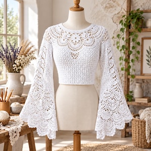 May include: A white long-sleeve crochet top featuring a round neckline with intricate floral and fan-shaped patterns. The garment has wide, flared bell sleeves and a cropped length, showcasing detailed needlework throughout the bodice and arms.
