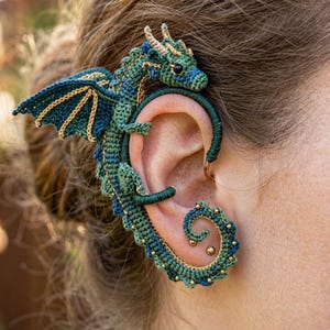 May include: A green and blue crocheted dragon ear cuff featuring intricate threadwork, small gold-colored accents, and a detailed wing design that wraps around the outer edge of the ear, secured by a thin wire frame structure.