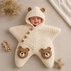 May include: A cream-colored, star-shaped crochet baby bunting with a hood featuring two small bear ears. The garment has four brown buttons along the diagonal front and two brown bear face appliques on the lower points of the star shape.