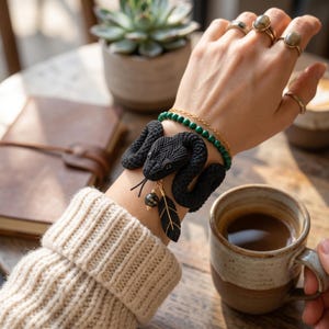 May include: A black macrame snake bracelet featuring a textured, coiled design with a small dangling charm shaped like a feather and a round bead. The bracelet is worn alongside a thin gold chain and a strand of small green malachite beads.
