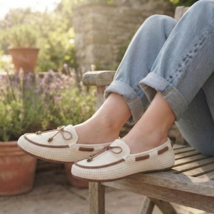 Puede incluir: Un par de mocasines de ganchillo color crema con ribete marrón en los bordes y un lazo decorativo marrón en la parte superior. Los zapatos tienen un aspecto tejido y texturizado con punta redondeada y diseño bajo, usados con pantalones vaqueros azules.