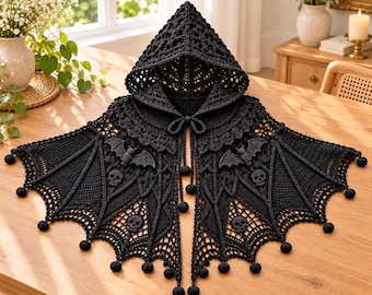May include: A black crocheted hooded cape featuring an intricate web-like pattern, decorative bat motifs, and skull accents. The garment includes a drawstring closure with pom-pom ends and a scalloped hemline finished with small black pom-poms.