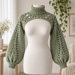 Puede incluir: Un bolero de ganchillo color verde salvia con cuello alto de canalé y mangas largas y voluminosas tipo obispo. La prenda presenta un intrincado patrón de encaje calado en los hombros y la parte superior del pecho, con puños de canalé.