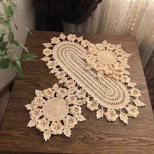May include: A set of three beige crochet doilies featuring an intricate oval runner and two matching circular mats. Each piece is crafted with a central solid disc surrounded by a border of three-dimensional floral motifs and pointed leaf accents.