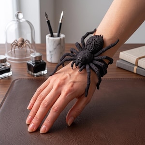 May include: A black spider-shaped bracelet featuring a textured, detailed body and eight long, curved legs that wrap around the wrist. The spider has small, dark, bead-like eyes and a matte finish, creating a realistic appearance on the wrist.