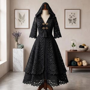 Könnte beinhalten: Ein schwarzes Kleid mit Kapuze und aufwendigen Spitzenmustern aus Blatt- und Blumenmotiven. Das Kleidungsstück hat dreiviertellange Ärmel, einen V-Ausschnitt und zwei goldfarbene, dekorative Knebelverschlüsse mit Quasten auf der Brust.