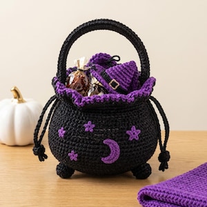Könnte beinhalten: Ein schwarzer, gehäkelter Kesselkorb mit violettem Rand, verziert mit violetten Stern- und Mondsichel-Applikationen. Der Korb hat einen stabilen Henkel, drei kleine Füße und enthält verpackte Süßigkeiten sowie einen kleinen Hexenhut.
