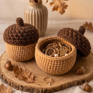 May include: Two crocheted acorn-shaped containers with dark brown caps and light tan bases. One container is closed, while the other is open, revealing gold-toned jewelry including rings and a leaf-shaped pendant inside. Two rings rest nearby.