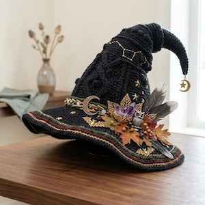 May include: A black crocheted pointed hat featuring gold star embroidery, a crescent moon charm, purple crystal clusters, an owl figurine, dark feathers, and autumn leaf accents. A small gold moon and star charm hangs from the tip of the hat.