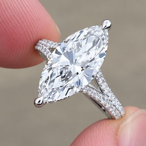Op de afbeelding: Een zilverkleurige ring met een grote marquise-geslepen middensteen die door twee pootjes wordt vastgehouden. De ringband is gespleten in twee delen, beide bezet met kleine, ronde, heldere steentjes die langs de zijkanten van de zetting lopen.