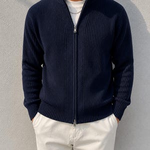 Puede incluir: Un suéter de punto acanalado azul marino con cremallera frontal completa, cuello alto y mangas largas con puños acanalados. El suéter se lleva sobre una camiseta blanca lisa y se combina con pantalones blancos, mostrando un diseño texturizado.