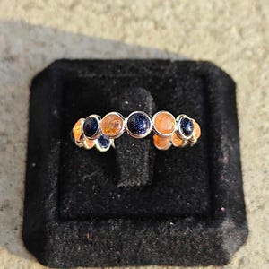 Op de afbeelding: Een zilverkleurige ring met een rij afwisselende ronde cabochonstenen. De stenen bestaan uit donkerblauwe glitterende zandsteen en doorschijnende oranje zonnesteen, elk gezet in een gladde metalen zetting langs de band van de ring.