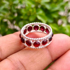 Op de afbeelding: Een zilveren ring met een gedraaid touwontwerp, bezet met een doorlopende rij ronde, dieprode granaatedelstenen. De metalen band heeft een gepolijste afwerking en de stenen zijn geplaatst in individuele ronde zettingen rondom de hele ring.