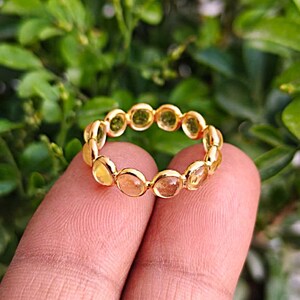 Könnte beinhalten: Ein goldener Ring mit einer durchgehenden Reihe runder, gelber Edelsteine in Zargenfassung. Der Ring besteht aus poliertem, goldfarbenem Metall, wobei jeder einzelne Stein in einer kreisförmigen Fassung sitzt, die miteinander verbunden sind.