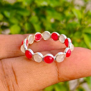Può includere: Un anello a fascia in argento con un motivo alternato di pietre rotonde, lisce e opache di colore rosso e pietre bianche traslucide, incastonate in castoni metallici individuali che formano un design circolare continuo sull'anello.
