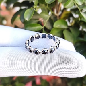 May include: A silver ring featuring a continuous band of small, round, polished black stones set in individual metal bezels. The stones are dark and reflective, arranged in a circular pattern around the band, creating a uniform and linked appearance.