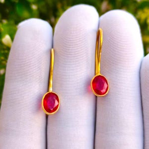 Könnte beinhalten: Ein Paar goldene Ohrhänger mit ovalen, facettierten roten Edelsteinen. Jeder Ohrring besteht aus einem dünnen, gebogenen Golddraht-Haken, der an einem in einer Fassung sitzenden, leuchtend roten Stein befestigt ist, mit polierter Metalloberfläche.