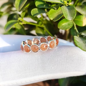 Op de afbeelding: Een zilveren ring met een doorlopende band van ronde, gefacetteerde zonnesteen edelstenen. Elke oranjebruine steen is gezet in een gepolijste zilveren zetting, wat een uniform cirkelvormig ontwerp creëert dat de hele ring omringt met textuur.